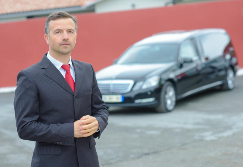 alt="funeral director with hearse in background"