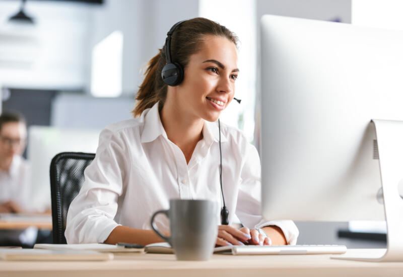 Happy cheerful business woman in office callcenter working with computer wearing headphones
